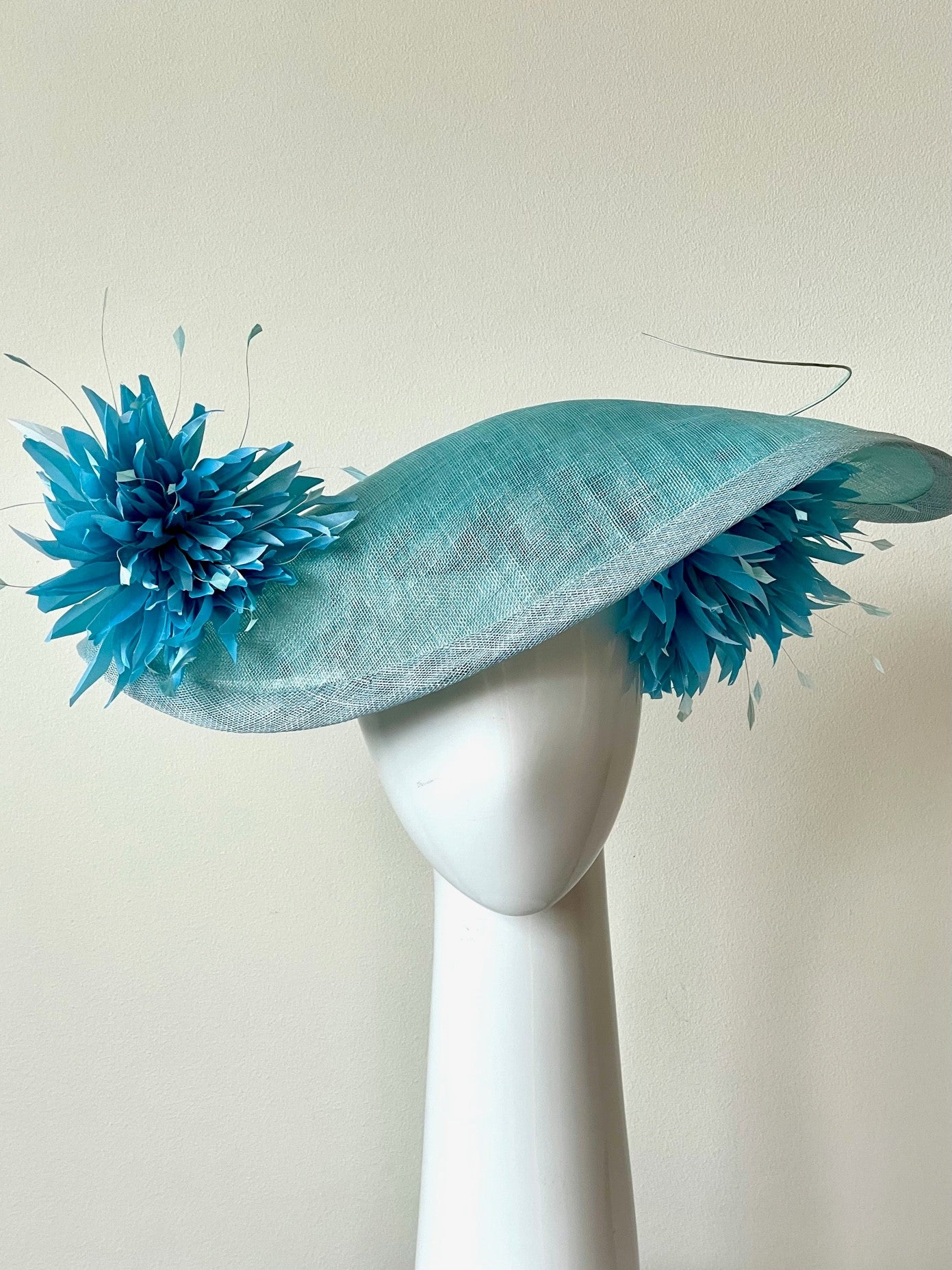 The Pale Blue Asymmetric Picture Hat features a curved oval platter hat and is finished with pom pom chrysanthemums under and on top of the brim.