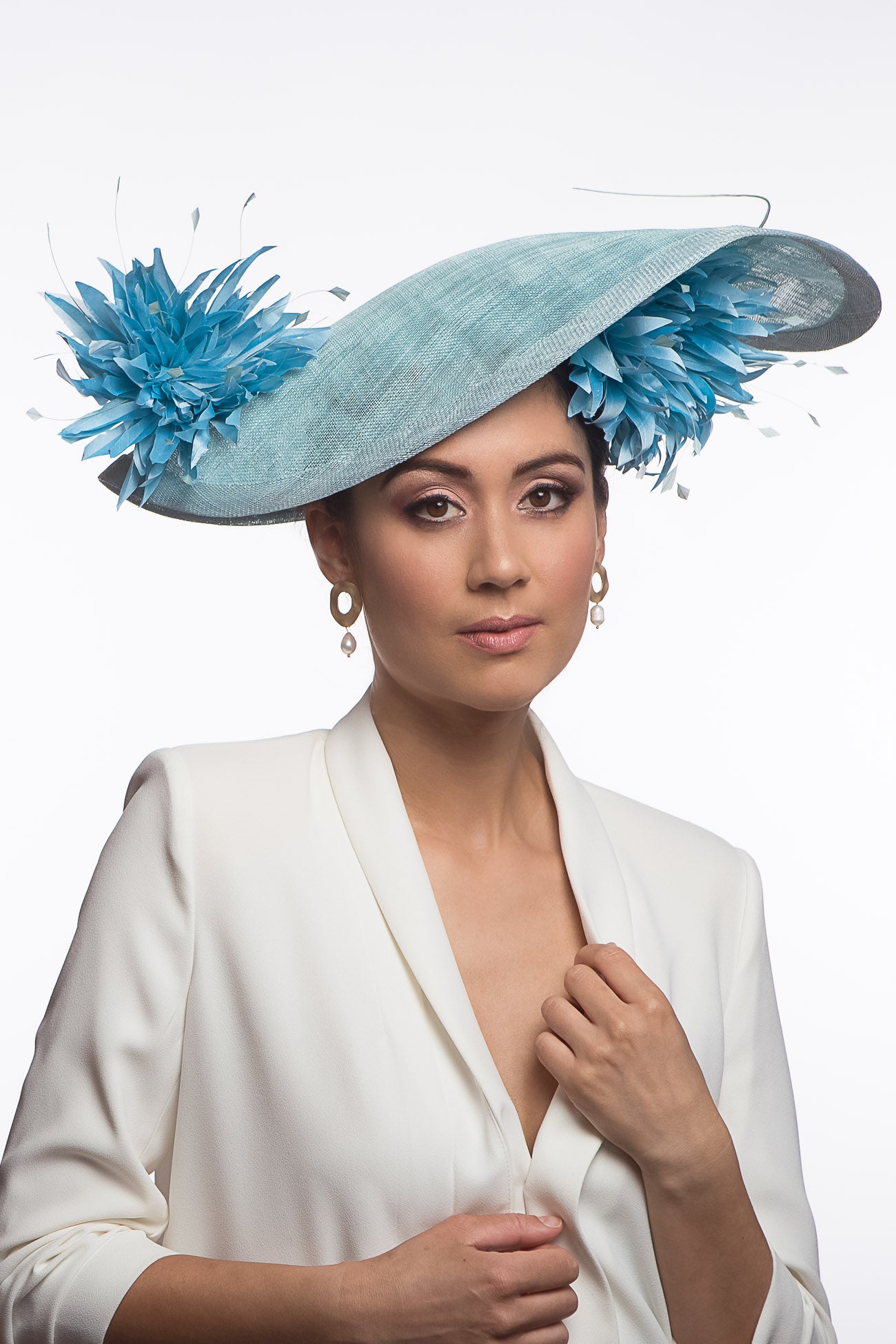 The Pale Blue Asymmetric Picture Hat features a curved oval platter hat and is finished with pom pom chrysanthemums under and on top of the brim.