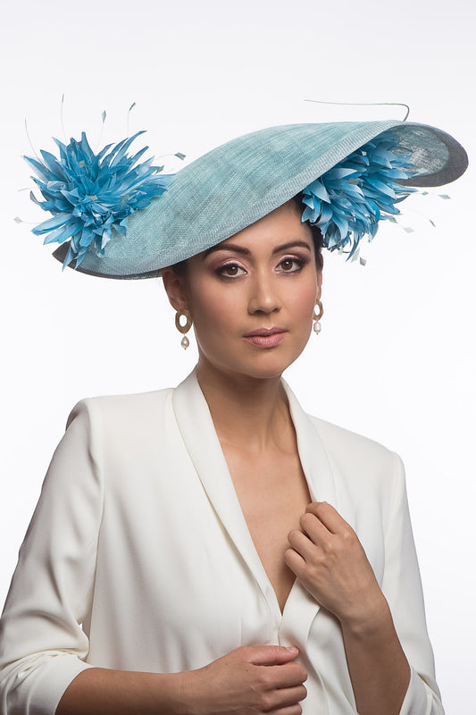 The Pale Blue Asymmetric Picture Hat features a curved oval platter hat and is finished with pom pom chrysanthemums under and on top of the brim.