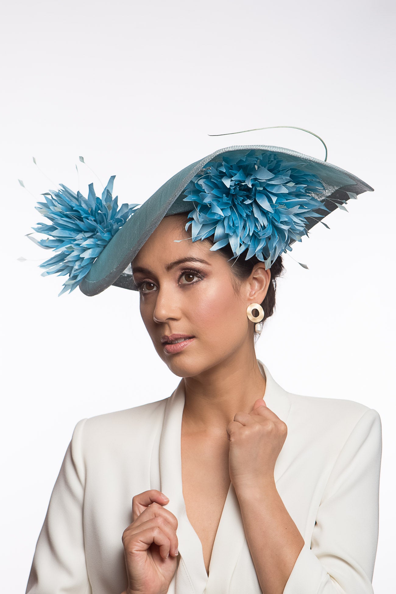 The Pale Blue Asymmetric Picture Hat features a curved oval platter hat and is finished with pom pom chrysanthemums under and on top of the brim.