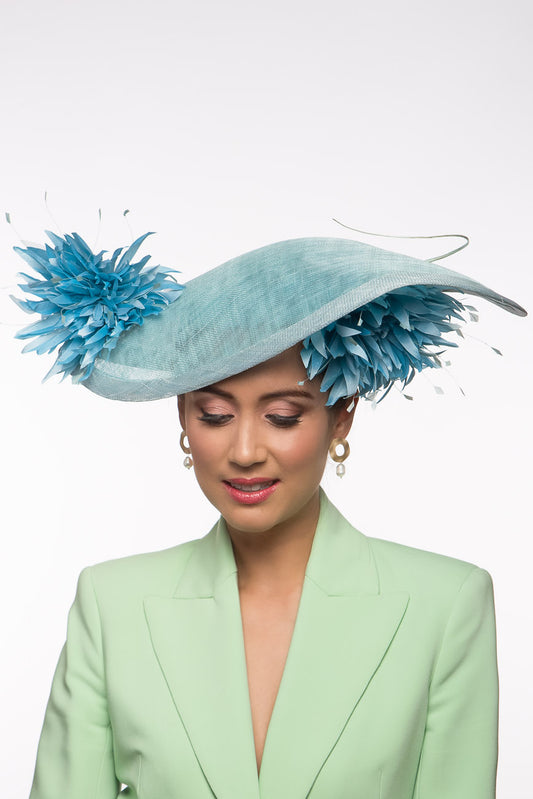 The Pale Blue Asymmetric Picture Hat features a curved oval platter hat and is finished with pom pom chrysanthemums under and on top of the brim. 