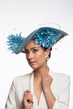 Load image into Gallery viewer, The Pale Blue Asymmetric Picture Hat features a curved oval platter hat and is finished with pom pom chrysanthemums under and on top of the brim. 