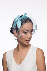 The Veiled Feather Leaf Headband in Blues features a birdcage veil adorned with feather leaves. The sky blue feather leaves are arranged over the pale blue ones